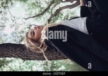 Die blonde Frau in Dreadlocks lehnt sich im Wald an einen Baum, trägt Blazer, T-Shirt und Hosen mit geschlossenen Augen und leicht geöffnetem Mund. Stockfoto