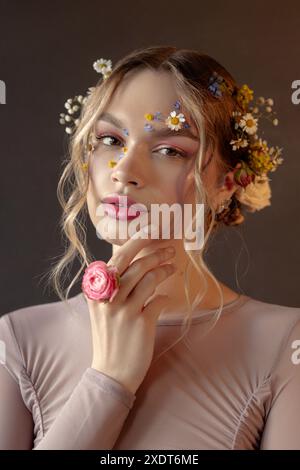 Eine Frau mit blonden Haaren und Blumen im Haar und im Gesicht, die ein hellrosa Langarmshirt trägt, schaut direkt in die Kamera. Stockfoto