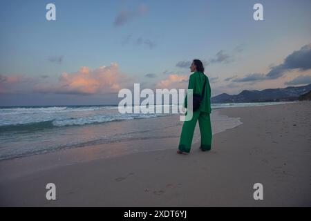 Frau in grüner Kleidung läuft an einem Sandstrand entlang, während die Sonne über dem Meer untergeht. Stockfoto