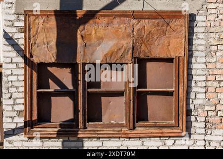 Altes braunes Holzfenster mit rostigem Blech an weißer Ziegelwand unter direktem Sonnenlicht Stockfoto