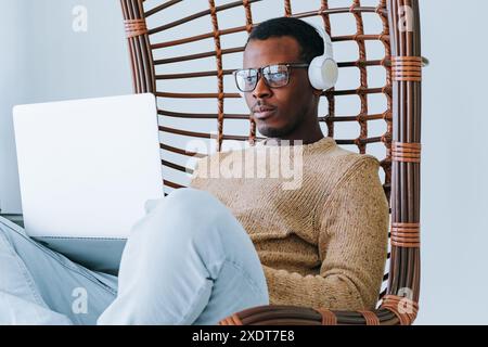 Ein afroamerikanischer Mann mit Brille und Kopfhörern arbeitet an seinem Laptop, während er bequem im Korbstuhl sitzt, konzentriert und entspannt. Modernes und stilvolles Büro mit komfortablen Arbeitsbereichen. Stockfoto