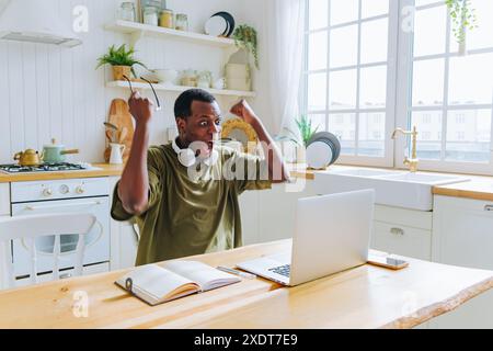 Afroamerikaner in der Küche, Kopfhörer um den Hals, feiert mit erhobenen Armen, während er auf seinen Laptop blickt, was einen Moment des Erfolgs anzeigt. Aufgeregter Mann, der Erfolg feiert. Stockfoto