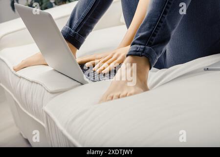 Die junge Frau sitzt bequem auf einem weißen Sofa in ihrem modernen Wohnzimmer und arbeitet mit den Füßen nach oben an einem Laptop, was einen gemütlichen, freiberuflichen Lebensstil widerspiegelt Stockfoto