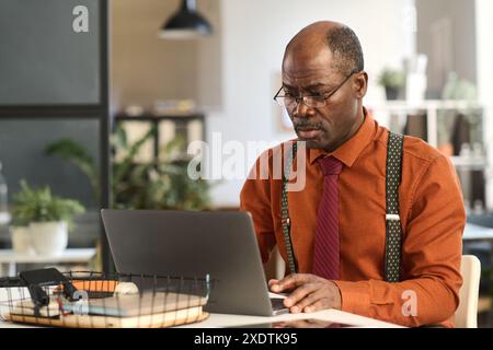 Porträt eines afroamerikanischen Seniorenmannes mit Laptop während der Arbeit im Büro und in alten Schuloutfits mit Hosenträgern, Kopierraum Stockfoto