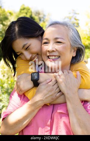 Asiatische Frau umarmt ältere Mutter von hinten, beide lächeln im Freien Stockfoto