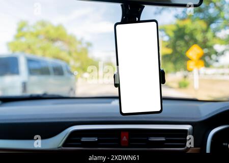 Ein leeres Smartphone mit weißem Bildschirm auf einem Armaturenbrett im Auto mit einer malerischen Straße, die durch die Windschutzscheibe sichtbar ist. Smartphone mit leerem Bildschirm in der Windschutzscheibe des Autos Stockfoto