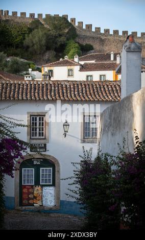 Rua de São Teotónio, Óbidos, Portugal. Eine enge Kopfsteinpflasterstraße führt zu einem Restaurant in einem traditionellen, zweistöckigen Dach mit Terrakotta-Fliesen und weiß getünchten Gebäude. Der nördliche Teil der Kalksteinmauer, die das Dorf im Hintergrund umgibt. Stockfoto