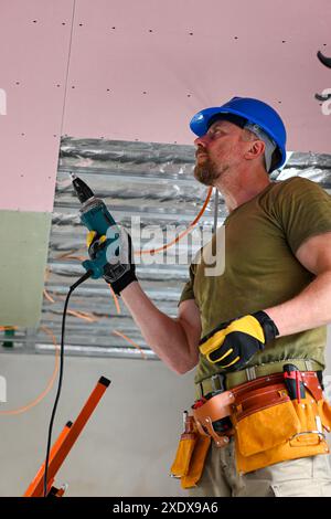 Bauarbeiter tragen Werkzeuggürtel und Sicherheitsausrüstung mit Bohrmaschine, während Trockenbauwände an der Decke montiert werden. Stockfoto