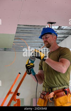 Bauarbeiter tragen Werkzeuggürtel und Sicherheitsausrüstung mit Bohrmaschine, während Trockenbauwände an der Decke montiert werden. Stockfoto