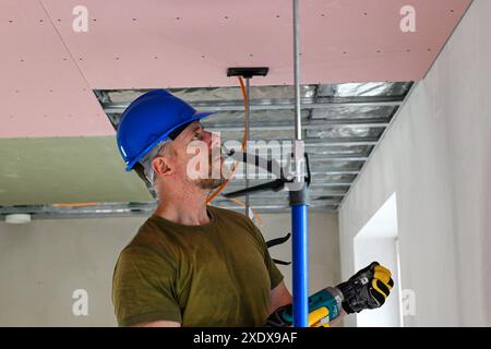 Bauarbeiter tragen Werkzeuggürtel und Sicherheitsausrüstung mit Bohrmaschine, während Trockenbauwände an der Decke montiert werden. Stockfoto