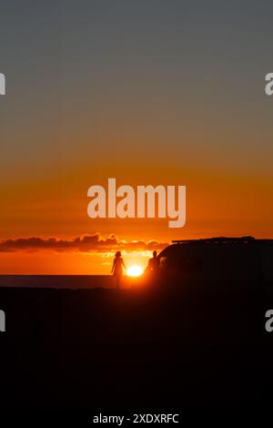 Ein Paar und ihr Fahrzeug haben eine Silhouette vor einem lebhaften Sonnenuntergang. Die untergehende Sonne färbt den Himmel in feurigen Orange- und Rottönen und wirft lange Schatten Stockfoto