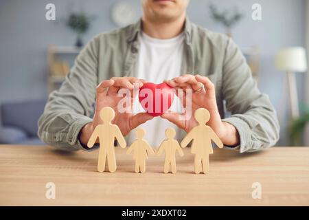 Mann, der ein rotes Herz über kleinen Holzfiguren von Mutter, Vater und Kindern hält Stockfoto