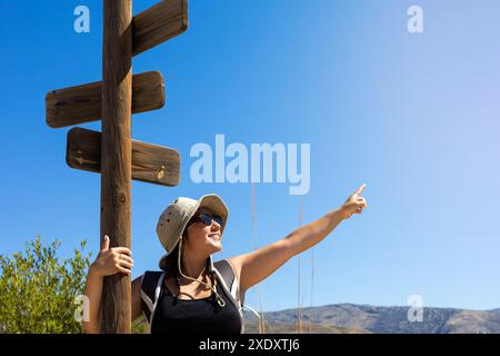 Lächelnde Frau mit Hut und Sonnenbrille zeigt begeistert auf einen Weg während einer Wanderung unter einem klaren blauen Himmel. Stockfoto