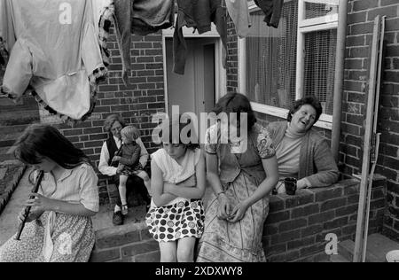 Das tägliche Leben in einem Kohlebergbaudorf in den 1970er Jahren Yorkshire, Großbritannien. Kinder von Bergleuten sitzen auf der Gartenwand, das Mädchen spielt eine Blockflöte und ihre Mutter unterhält sich bei einer Tasse Tee mit ihrem Nachbarn. Vater hat eines seiner Kinder auf dem Schoß. Die kürzlich gewaschenen Kleider hängen zwischen den Häusern, um sie zu trocknen. South Kirkby, Yorkshire England 1979. HOMER SYKES Stockfoto