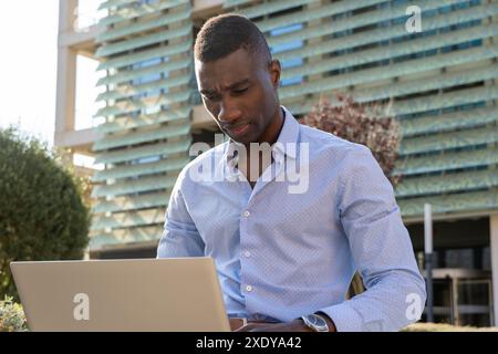Junger afrikanischer Geschäftsmann, der mit Laptop im Freien arbeitet, im Finanzviertel, in der Wirtschaft, in der Technologie, in der Fernarbeit, in der Stadt Stockfoto