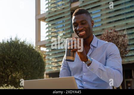 Lächelnder junger afroamerikanischer Geschäftsmann mit Laptop und Kaffee, der draußen im Finanzviertel, in der Geschäftswelt, in der Technologie, in der Stadt arbeitet. Stockfoto