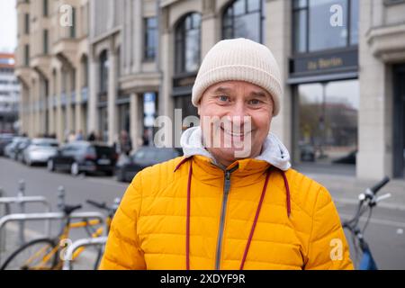 Reifer charismatischer Mann, 65 in hellgelber Pufferjacke und Mütze, Senior lächelt auf der städtischen Straße mit historischen Gebäuden im Hintergrund, Spaziergänge in E Stockfoto