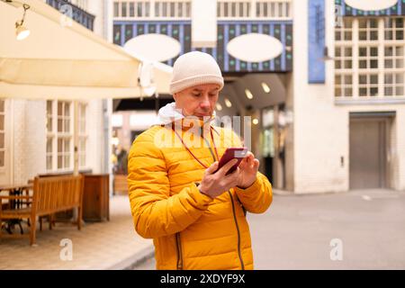 Seniorenmann in knallgelber Jacke mit Smartphone auf der städtischen Straße. Moderne Technik trifft auf historische Architektur in der Stadtszene, Spaziergänge in Europa Stockfoto