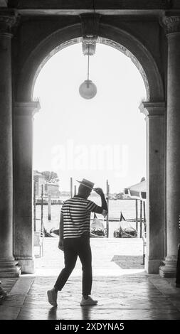 Venedig, Italien - 30. Juni 20220 Gondoliere, der von der Kamera unter einem Bogengang mit Gondeln im Hintergrund weggeht Stockfoto