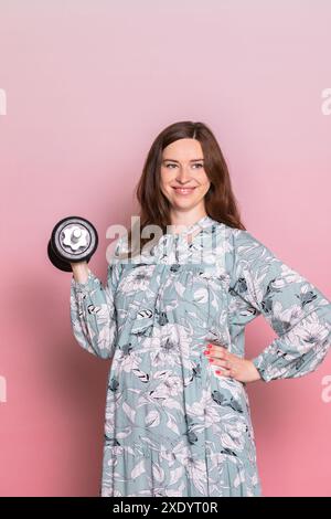 Glückliche junge schwangere Frau trägt blaue Kleid tun Übung heben Hantel. Studio shot auf rosa Hintergrund. Fitness und gesunde Lebensweise während der Pr Stockfoto