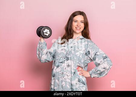 Glückliche junge schwangere Frau trägt blaue Kleid tun Übung heben Hantel. Studio shot auf rosa Hintergrund. Fitness und gesunde Lebensweise während der Pr Stockfoto