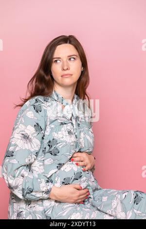 Nachdenkliche junge schwangere Frau im Blumenkleid, die Hände auf dem Bauch hält. Studio-Aufnahme auf rosa Hintergrund mit Kopierraum Stockfoto