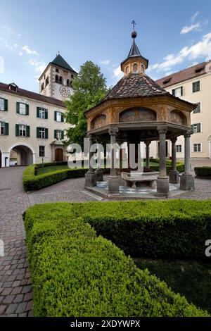 Novacella Abbey: Der „Wunderbrunnen“ im Innenhof und der Glockenturm mit der Uhr. Varna, Südtirol, Italien. Stockfoto