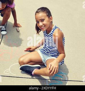 Mädchen, Kinder und Lächeln auf dem Boden mit Kreide auf Beton für Porträt, Spiel und Kreativität im Sommer. Kind, Zeichnen und Kunst bei Sonnenschein, Bürgersteig und Stockfoto