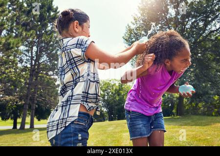 Wasserballons, glückliche und Mädchen im Park mit Freiheit, Spaß und Verbundenheit beim Lernen in der Natur. Lachen, Spritzen und Freunde mit Flüssigkeit Stockfoto