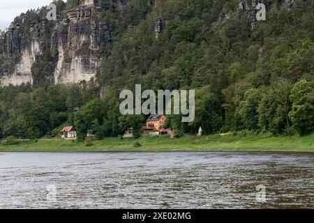 Eine Bergkette mit einem Fluss, der durch sie fließt, und einem Haus am Ufer. Das Haus ist orange und hat ein Steindach. Das Wasser ist ruhig und die Bäume A Stockfoto