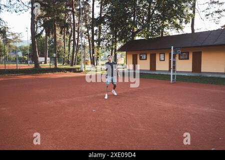 Ein schwarzhaariger Athlet mit einem Lächeln im Gesicht spielt Badminton auf einem Tonplatz. Begeisterung für den Amateursport. Lernen neuer Bewegungsfähigkeiten. Stockfoto