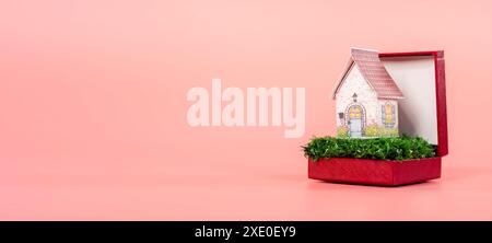 Ein kleines Haus sitzt in einem roten Kasten auf rosa Hintergrund. Das Haus ist mit Gras und Blumen dekoriert, was ihm eine gemütliche und einladende Atmosphäre verleiht. Den Stift Stockfoto