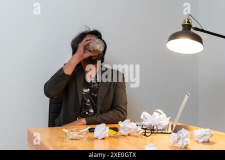 Das Konzept des Alkoholismus am Arbeitsplatz. Betrunkener Mann im Büro. Asiatischer Geschäftsmann versagt, Mann in Verzweiflung trinkt harten Alkohol, sitzt spät am Tisch Stockfoto