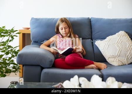 Das blonde Mädchen schläft ein und liest ein Buch auf einem blauen Sofa zu Hause Stockfoto