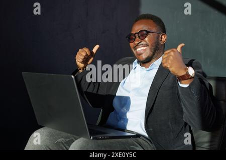 Glücklicher afrikanischer Geschäftsmann mit Laptop mit Daumen nach oben ausgezeichnetem Schild auf dunklem Hintergrund, erfolgreicher Geschäftsmann Stockfoto