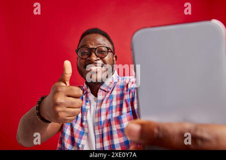 Ein junger Afroamerikaner freut sich, ein Telefon in der Hand zu halten und zeigt auf rotem Hintergrund eine Daumen-nach-oben-Geste, der Blogger sendet live Stockfoto