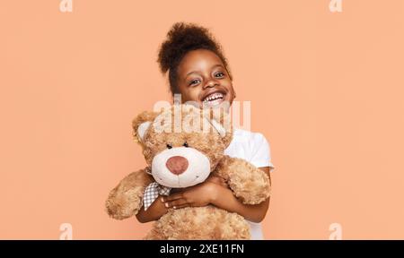 Kleines Mädchen, das Teddybär hält und vor Peach-Hintergrund lächelt Stockfoto