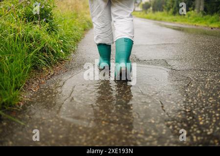 Frau mit Regengummistiefeln, die beim Herbstregen mit Wasserspritzern und Tropfen in die Pfütze laufen und springen. Stockfoto