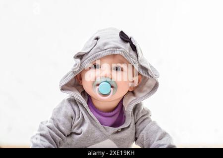 Fröhliches Mädchen in einer grauen Hasenjacke krabbelt glücklich auf den Boden und erforscht drinnen mit einem Schnuller im Mund Stockfoto