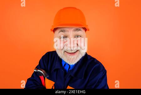 Lächelnder bärtiger Mann in Bauhelm und Uniform mit Hammer. Bauindustrie. Nahaufnahme Porträt eines hübschen Baumeisters, Reparaturmannes, Zimmermanns Stockfoto