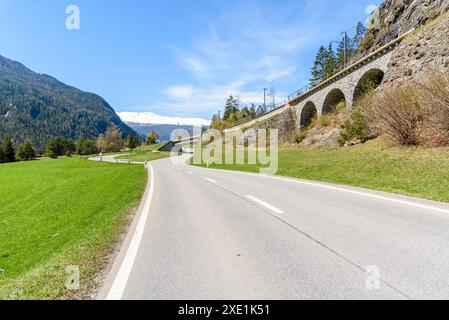 Einsame kurvige Straße durch ein Tal, die an einem klaren Frühlingstag unter einer Eisenbahn in den Schweizer Alpen vorbeiführt Stockfoto