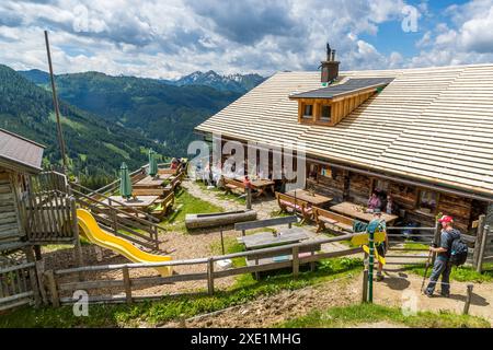Die Ellmaualm, auf einer Höhe von 1.794 Metern im Großarltal, Salzburg, Österreich, begrüßt Wanderer mit einem frisch schindelnbedeckten Dach, Solarpaneelen und einer Terrasse voller Gäste an Holztischen. Zu linker Hand der Hütte, die von der Familie Hettegger betrieben wird, stehen ein gelber Rutsche und ein Kinderspielplatz, während ein Wanderer mit rotem Hut von rechts herankommt. Stockfoto