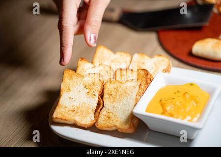 Eine Hand greift nach gerösteten Brotscheiben, serviert mit einer Schüssel Käse Dip auf einer weißen Platte. Stockfoto