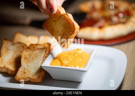 Nahaufnahme einer Hand, die geröstetes Brot in eine Schüssel Käsesauce taucht, mit mehr gerösteten Brotscheiben auf einem Teller und einer verschwommenen Pizza im Hintergrund. Stockfoto