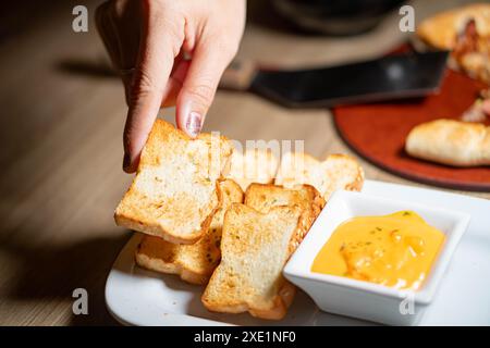 Eine Hand greift nach gerösteten Brotscheiben, serviert mit einer Schüssel Käse Dip auf einer weißen Platte. Stockfoto