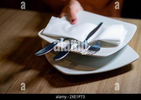 Nahaufnahme einer Hand, die einen Stapel weißer Teller mit Serviette und Besteck auf einen Holztisch legt. Stockfoto