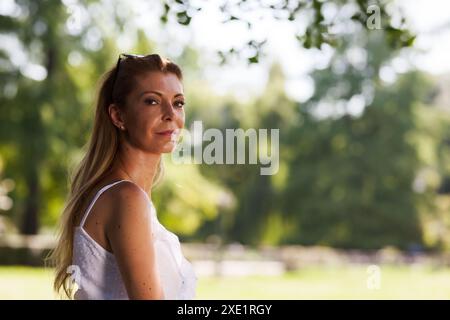 Eine Frau mittleren Alters in einem weißen ärmellosen Kleid steht selbstbewusst in einem üppigen Park, wo das Sonnenlicht durch die Bäume gefiltert wird. Stockfoto