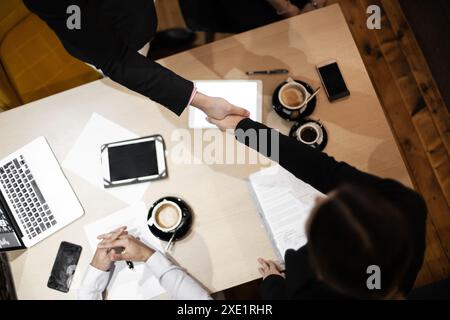 Draufsicht von zwei Geschäftsleuten, die bei einer Besprechung die Hand schütteln. Laptops, Smartphones und Kaffeetassen sind auf dem Tisch sichtbar. Stockfoto