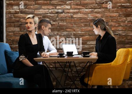 Drei junge Fachkräfte nehmen an einem produktiven Meeting in einem Café Teil, bei dem Laptops und Kaffee auf dem Tisch stehen und Teamarbeit und Geschäftskommunikation präsentieren. Stockfoto