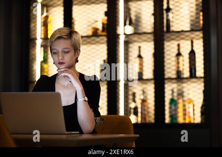 Eine junge blonde Frau in einem schwarzen Outfit, die sich in einer Bar mit einem wunderschön beleuchteten Flaschenregal hinter sich auf ihren Laptop konzentriert. Stockfoto